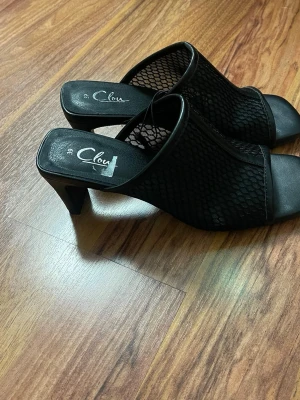 Svarta mesh-pumps från Clou - Snygga svarta pumps från Clou med öppen tå och klack. Ovandelen är i mesh-tyg med nätmönster och detaljer i konstläder. Skorna har slip-in-modell och en stilren, modern look som passar till många outfits.