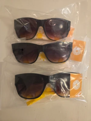 Svarta solglasögon Sunshine - Snygga svarta solglasögon från Sunshine med bred båge och rektangulär form. Glasen är mörka med en lätt toning som går från svart till brunt. Perfekta för soliga dagar och levereras med en gul förvaringspåse.