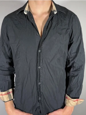 Svart skjorta från Burberry - Stilren svart skjorta från Burberry med klassiskt rutigt mönster på insidan av kragen och manschetterna. Skjortan har knappar framtill och lång ärm. Perfekt för dig som gillar en tidlös look med en twist.