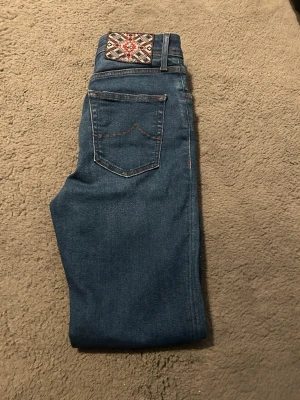 Mörkblå jeans med broderad patch - Snygga mörkblå jeans från cohën med en unik broderad patch i rött, vitt och svart på linningen. Klassisk femficksmodell med kontrastsömmar och rak passform. Perfekta för dig som vill ha något extra detaljerat i garderoben. Tror de är dam modell men passar även för killar