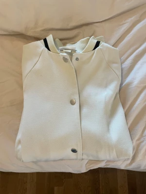 Vit jacka med svarta detaljer - Snygg vit bomberjacka med ribbade muddar och svarta ränder vid krage, ärmslut och nederkant. Jackan har tryckknappar framtill och två sidofickor. Oversized passform och sportig look, perfekt för dig som gillar streetwear.