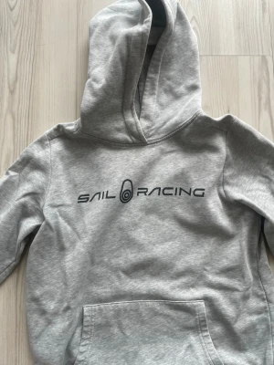 Grå hoodie från Sail Racing - Snygg grå hoodie från Sail Racing med svart logga tryckt på bröstet. Hoodien har en stor känguruficka framtill och en klassisk huva. Tillverkad i mjukt bomullsmaterial som känns skönt mot huden. Perfekt för en avslappnad och sportig stil.