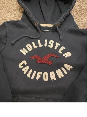 Mörk blå hoodie från Hollister - Mörkblå hoodie från Hollister med broderad text 'Hollister California' och röd logga på bröstet. Klassisk känguruficka framtill, ribbade muddar och justerbar huva med snören. Mjuk bomullsblandning som känns skön mot huden.