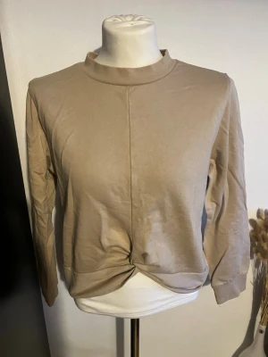 Beige långärmad topp med twist - Snygg beige långärmad topp med twistad detalj framtill. Toppen har en högre krage och är tillverkad i mjukt material som ger en avslappnad men ändå stilren look. Perfekt att matcha med jeans eller kjol för en trendig outfit.