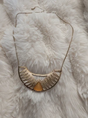 Guldigt halsband med geometrisk detalj - Unikt halsband i guldfärgad metall med en halvcirkelformad, strukturerad hängare och en triangulär gul sten i mitten. Kedjan är tunn och smidig, och designen har en modern, bohemisk känsla som sticker ut.