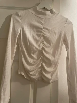 Vit ribbad topp med rynk framtill - Trendig vit långärmad topp med ribbad struktur och rynkdetalj längs magen. Hög hals och figurnära passform ger en snygg siluett. Perfekt att styla med jeans eller kjol för en cool look.