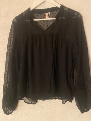 Svart blus - Svart blus från Only med transparenta långärmar och diskret prickigt mönster. Blusen har v-ringning med knytband och spetsdetaljer vid axlarna. Perfekt för dig som gillar en stilren men ändå detaljrik look.