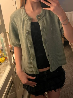 Grön kortärmad kofta med knappar - Snygg ljusgrön kofta med korta ärmar och stora ribbade detaljer vid krage, knappslå och nederkant. Koftan har stora matchande knappar framtill och är gjord i ett mjukt stickat material. Perfekt att slänga över ett linne för en chill look.