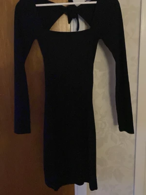 Svart långärmad klänning med knyt - Stilren svart klänning med lång ärm och snygg knytning i ryggen. Modellen är kroppsnära och kort.