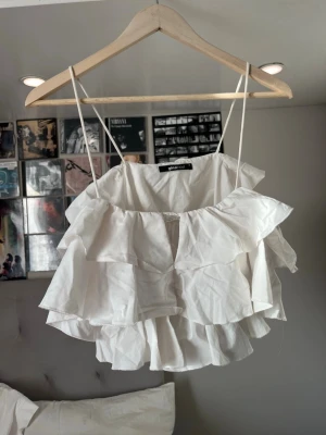 Vit volangtop från Gina Tricot - Supersöt vit linnetopp från Gina Tricot med tunna axelband och flera lager volanger. Perfekt för dig som gillar en luftig och lekfull stil. Toppen har en lös passform och är tillverkad i ett lätt bomullstyg.