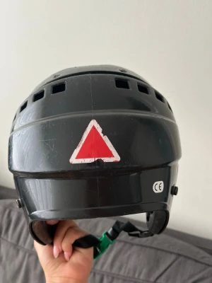 Svart hjälm från Reebok - Säljer en svart hjälm från Reebok med ventilationshål och justerbart hakband. Hjälmen har vita och röda triangulära dekaler och CE-märkning på baksidan. Perfekt för dig som vill ha en enkel och funktionell hjälm till olika aktiviteter.