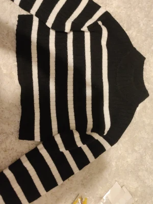 Svartvit randig stickad tröja - Säljer en svart stickad tröja med vita horisontella ränder. Tröjan har en croppad passform, långa ärmar och ribbstickad halsringning. Perfekt för dig som gillar en klassisk men trendig look.