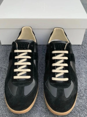 Svarta sneakers från Maison Margiela - Stilrena svarta sneakers från Maison Margiela med ovandel i mocka och läder, vita snören och beige yttersula i gummi. Klassisk low-top siluett och diskret logga på plösen. Perfekta för dig som gillar minimalistisk och tidlös design.