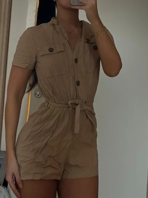 Beige kortärmad jumpsuit med knappar - Snygg beige jumpsuit med korta ärmar, knappar framtill och två bröstfickor. Midjan har ett matchande knytskärp som ger en schysst siluett. Materialet ser ut att vara mjukt och lätt, perfekt för varma dagar. Enkel och trendig design som passar till mycket. behöver strykas 