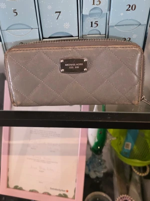 Grå quiltad plånbok Michael Kors - Snygg grå plånbok från Michael Kors med quiltat mönster och silverfärgad metallbricka framtill. Plånboken har dragkedja runtom och är tillverkad i ett glansigt konstläder. Perfekt för dig som gillar stilrena accessoarer.