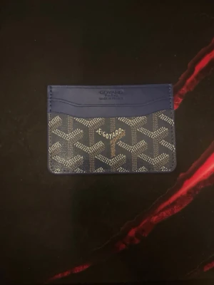 Blå kortfodral  - Snyggt blått kortfodral med klassiskt mönster i vitt och grått. Tillverkat i canvas och läder, har flera kortfack och den ikoniska loggan framtill. Perfekt för dig som vill ha en stilren och exklusiv accessoar i fickan.