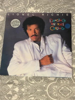 Lionel Richie Dancing on the Ceiling LP - Vinylskiva med albumet 'Dancing on the Ceiling' av Lionel Richie. Omslaget har färgglad text och visar en person i vit kostym. Inkluderar inneromslag med helkroppsbild. Perfekt för samlare av klassisk pop och soul.