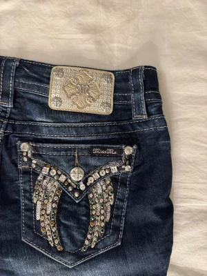 Miss Me jeansshorts med paljettdetaljer - Säljer ett par mörkblå jeansshorts från Miss Me med glittriga paljettdetaljer och nitar på bakfickan. Shortsen har låg midja och en snygg, dekorerad patch bak. Storlek 27 (motsvarande S)