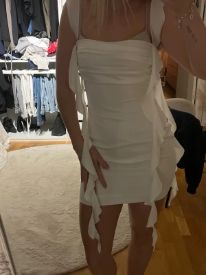 Vit kort klänning med volanger - Säljer en vit kort klänning med smala axelband och volangdetaljer längs sidorna. Klänningen har en figurnära passform och är perfekt för dig som vill sticka ut. Materialet känns mjukt och följsamt, troligen polyester eller liknande. Klänningen är från pappermayo aldrig använd och har prislappen kvar.