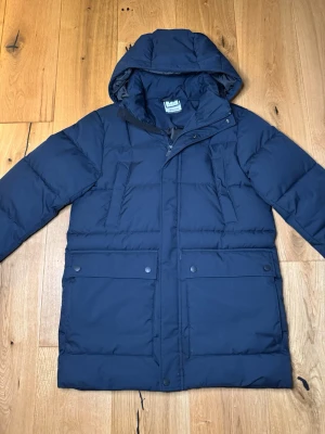 Mörkblå pufferjacka med huva - Snygg mörkblå pufferjacka från McKINLEY med huva och dragkedja framtill. Jackan har två stora fickor med lock och knappar samt två snedställda bröstfickor. Perfekt för kalla vinterdagar tack vare det vadderade materialet.