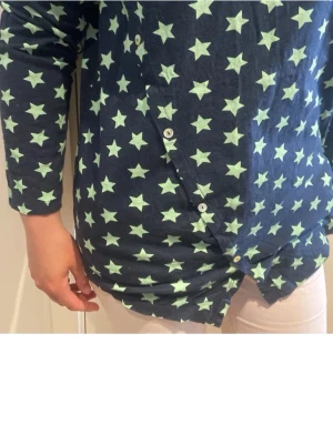 Mörkblå blus med stjärnmönster - Mörkblå blus med ljusgröna stjärnor över hela tyget. Blusen har rund halsringning, långa ärmar och dekorativa knappar framtill i omlottstil. Tillverkad i mjukt och bekvämt material, perfekt för en avslappnad look.