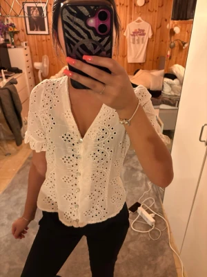 Vit blus med detaljer - Söt vit blus med hålmönster och volangkant vid ärmslut. Blusen har v-ringning, knappar framtill och är tillverkad i ett lätt, luftigt tyg som känns som bomull. Perfekt för en trendig och somrig look.