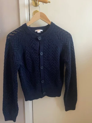 Marinblå stickad kofta med knappar - Snygg marinblå stickad kofta med genomgående knappar framtill. Koftan har ett hålmönster i zigzag, ribbade muddar och rund halsringning. Perfekt att slänga över en topp för en chill look.