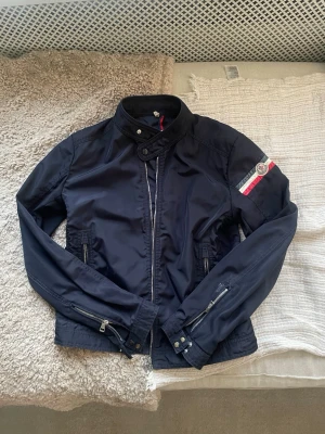 Mörkblå vindjacka från Moncler - Snygg mörkblå vindjacka från Moncler med ståkrage och dragkedja framtill. Jackan har två fickor med dragkedja, detaljerade muddar vid ärmslut och Moncler-logga på ärmen med röd, vit och blå rand. Perfekt för en sportig och trendig look. Storlek 3 motsvarar S/M. Öppen för byteshandel 