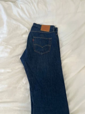 Mörkblå raka jeans från Levi's 501 - Säljer ett par klassiska mörkblå jeans från Levi's med raka ben och hög midja. Jeansen har två bakfickor med den ikoniska sömmen och en brun läderpatch i midjan. Tillverkade i slitstark denim med snygga kontrastsömmar.