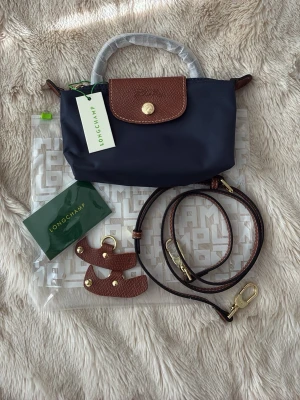 Mörkblå Longchamp Model depose väska - Snygg mörkblå handväska från Longchamp Le Pliage med bruna läderdetaljer och guldfärgad knapp. Väskan har avtagbar axelrem och klassisk vikbar design. Tillverkad i slitstark nylon med detaljer i präglat läder. Perfekt för dig som gillar stilrena accessoarer.