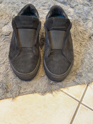 Axel arigato - Stilrena svarta slip-on sneakers från Axel Arigato i mocka. Skorna har en minimalistisk design utan snörning, med diskret logga på sidan och tjock svart sula. Perfekta för dig som gillar en clean och modern look.