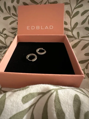 Silverfärgade hoops från Edblad - Snygga små hoops i silverfärg från Edblad. Örhängena är runda och har gnistrande stenar på framsidan som ger en elegant touch. Kommer i original ask. Perfekta för dig som gillar stilrena och moderna smycken.