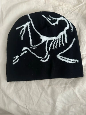 Mössa  - Säljer en svart mössa med en cool vit grafisk illustration som täcker framsidan. Mössan är mjuk och stretchig, perfekt för att hålla värmen och samtidigt ge en unik stil. Passar dig som vill sticka ut lite extra i vinter.