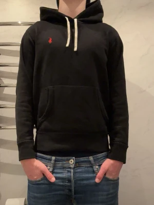 Svart hoodie från Ralph Lauren - Svart hoodie från Ralph Lauren med klassisk röd logga broderad på bröstet. Nypris 2000 använd fåtal gånger Hoodien har vit dragsko i huvan och en stor magficka framtill. Tillverkad i mjukt material och har ribbade muddar vid ärmar och nederkant. Perfekt för en avslappnad och stilren look.