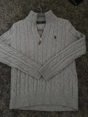 Grå Ralph lauren halfzip - Kabelstickad grå tröja från Polo Ralph Lauren med halv dragkedja och hög krage. Tröjan har långa ärmar, ribbade muddar och nederkant samt den klassiska broderade loggan på bröstet. Perfekt för lager på lager och en stilren look.