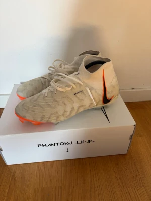 Nike Phantom Luna vita fotbollsskor - Säljer ett par Nike Phantom Luna fotbollsskor i vitt med grått mönster och orange sula. Skorna har hög ankelkrage och klassisk snörning. Stor svart-orange Nike swoosh på sidan. Perfekta för dig som vill ha stilrena och moderna skor på planen.