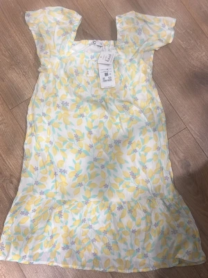 Blommig kortklänning i pastellfärger - Söt kortklänning med blommigt mönster i ljusgult, blått och vitt. Klänningen har volang nertill och korta ärmar med lätt puff. Perfekt för varma dagar och ger en fräsch sommarkänsla. Tillverkad i ett lätt och luftigt material.