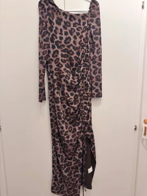Lång leopardmönstrad klänning - Säljer en långärmad klänning med leopardmönster i beige och svart. Klänningen har asymmetrisk halsringning, rynkade detaljer längs sidan och en slits nedtill. Materialet känns stretchigt och mjukt, troligen polyester. Perfekt för dig som vill sticka ut.