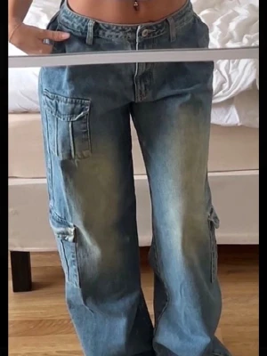 Blåa cargobyxor i denim - Säljer ett par blå cargobyxor i denim med bred passform och flera stora fickor på sidorna. Byxorna har en tvättad look med ljusare partier och klassisk cargostil. Perfekta för en avslappnad och trendig streetwear-outfit.