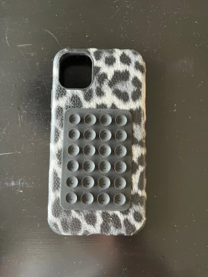 Leopardmönstrat mobilskal med pop it - Snyggt mobilskal med leopardmönster i svart, grått och vitt. På baksidan finns en svart pop it-panel som gör skalet extra kul och unikt. Skalet är gjort i ett mjukt material som skyddar mobilen och ger ett bra grepp. Passar dig som vill ha något trendigt och lekfullt.