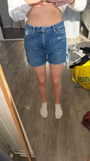 Blå denimshorts med råa kanter - Säljer ett par klassiska blå jeansshorts i denim med hög midja och råa, fransiga kanter. Shortsen har fem fickor, knappgylf och en avslappnad passform. Perfekta för sommardagar och enkel att matcha med olika toppar.