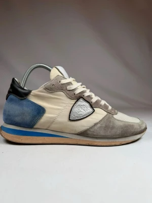 Philippe Model sneakers beige/blå - Snygga sneakers från Philippe Model i beige och grå mocka med blå och svart detalj på hälen. Skorna har vita meshpartier, vita skosnören och en blå rand på sulan. Klassisk låg modell med logga på sidan och plös. Storlek 41 men passar 42