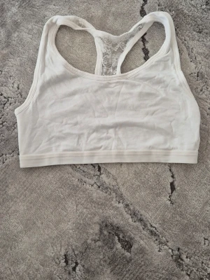 Vit sport-bh med spets från Esmara - Vit sport-bh från Esmara med brottarrygg och spetsdetalj i ryggen. Tillverkad i mjukt material för skön känsla och har bred resår nedtill. Enkel och stilren design som passar perfekt till träning eller chill.