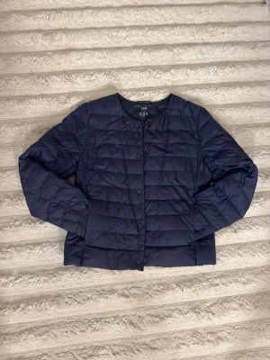 Mörkblå lättvikts pufferjacka Uniqlo - Snygg mörkblå pufferjacka från Uniqlo med rund halsringning och knäppning framtill. Jackan är quiltad i horisontella rutor och har en lätt, smidig passform som funkar perfekt till lager-på-lager. Tillverkad i tunt, vadderat material som håller värmen utan att kännas klumpig.