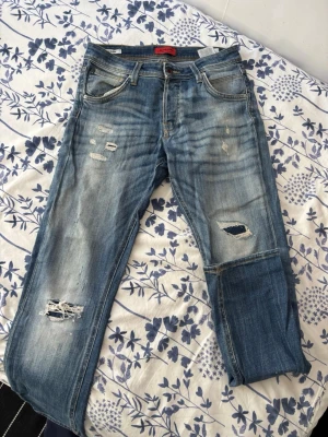 Blå slitna jeans från Jack & Jones - Säljer ett par blå jeans från Jack & Jones med slitningar och hål på benen för en cool, avslappnad look. Jeansen har klassisk femficksmodell, normal midja och raka ben. Perfekta för dig som gillar streetstyle och en trendig vibe.