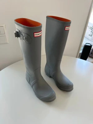 Grå höga gummistövlar från Hunter - Säljer ett par klassiska gråa höga gummistövlar från Hunter. Stövlarna har justerbart spänne på sidan och  Hunter-logga framtill. Storlek 38. Nyskick, aldrig använda. Upphämtas i Haninge eller Karlaplan. 