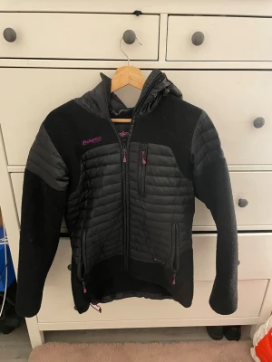 Svart vindjacka från Bergans - Snygg svart vindjacka från Bergans of Norway, modell Osen Lady Jacket. Jackan har quiltade partier, huva och dragkedjor med lila detaljer. Tillverkad i en mix av syntetmaterial och fleece för extra komfort och värme. Perfekt för blåsiga dagar och outdoor-äventyr.