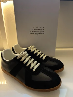 Maison Margiela svarta sneakers - Stilrena svarta sneakers från Maison Martin Margiela med vita snören och klassisk gummisula. Skorna har en låg silhuett och är tillverkade i en mix av läder och mocka för en lyxig känsla. Perfekta för dig som gillar minimalistisk och tidlös design.