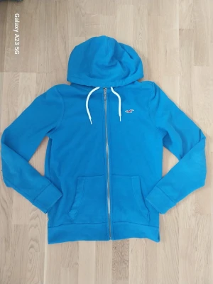 Blå zip hoodie från Hollister - Blå hoodie från Hollister med dragkedja framtill och vit dragsko i huvan. Klassisk Hollister-logga i rött på bröstet. Två fickor framtill och mjukt bomullsmaterial gör den perfekt för chill dagar.