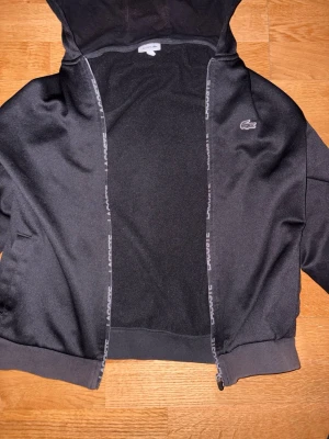 Svart zip hoodie från Lacoste - Snygg svart hoodie från Lacoste med dragkedja och huva. Klassisk krokodil-logga på bröstet och diskreta Lacoste-tryck längs dragkedjan. Tillverkad i mjukt och bekvämt material, perfekt för en chill och sportig look.